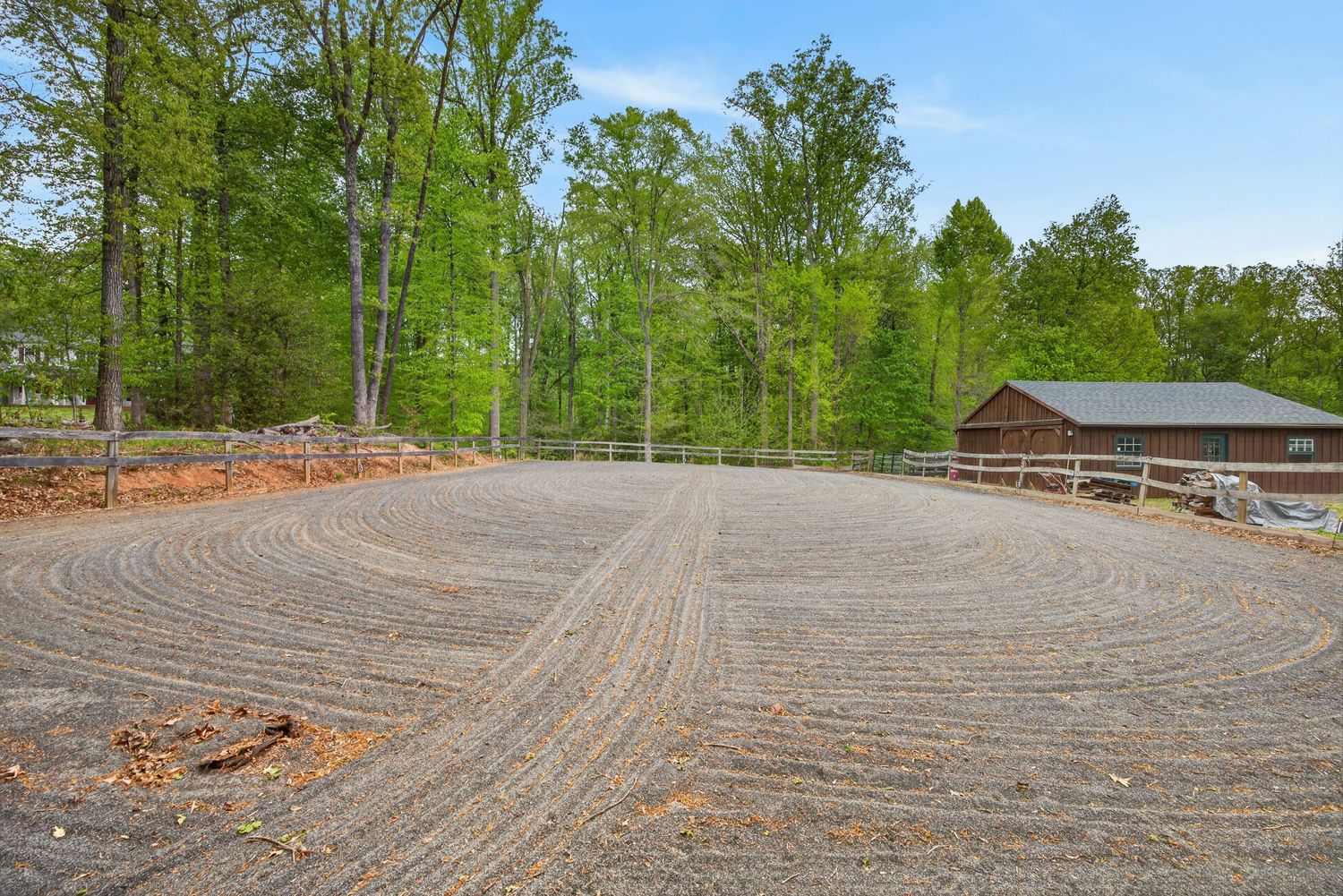Freshly graded gravel riding arena surrounded by lush green trees with a rustic barn visible in the background.