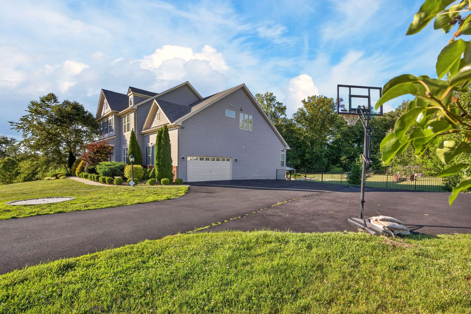 Suburban home with gray siding, attached garage, basketball hoop, and lush green lawn under a partly cloudy sky.