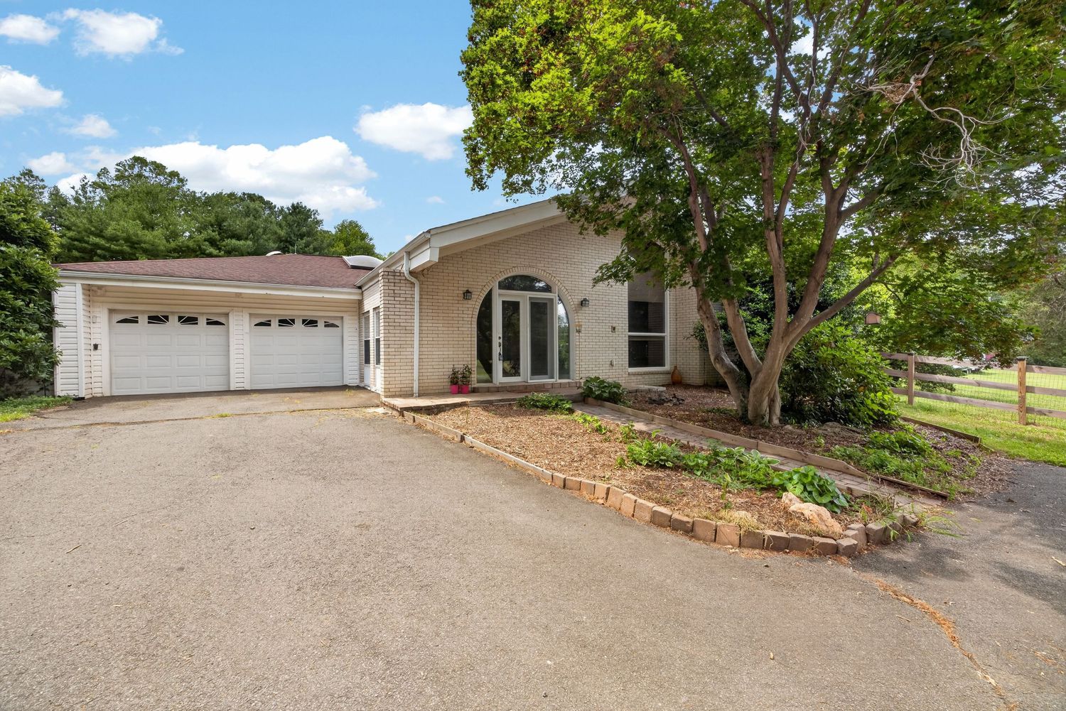 Ranch-style home with double garage, arched entry, lush trees, and a curved driveway surrounded by manicured landscaping.