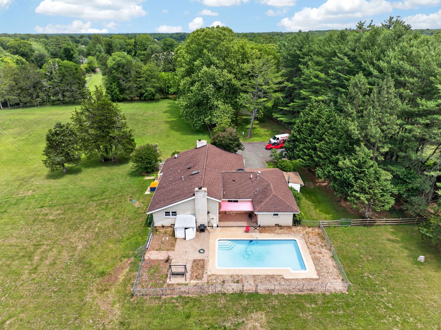 Aerial view of a suburban home with a backyard pool surrounded by lush green trees and open lawn.