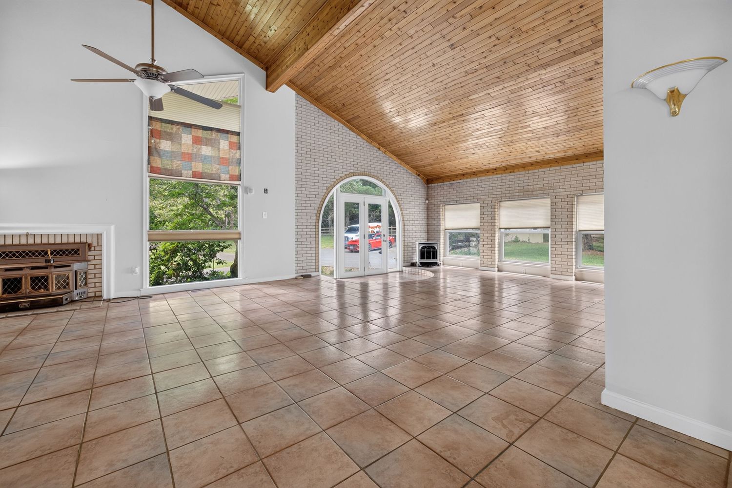 Spacious sunroom with high wood-paneled vaulted ceiling, tile floors, ceiling fan, arched doorway, and large windows with garden views.