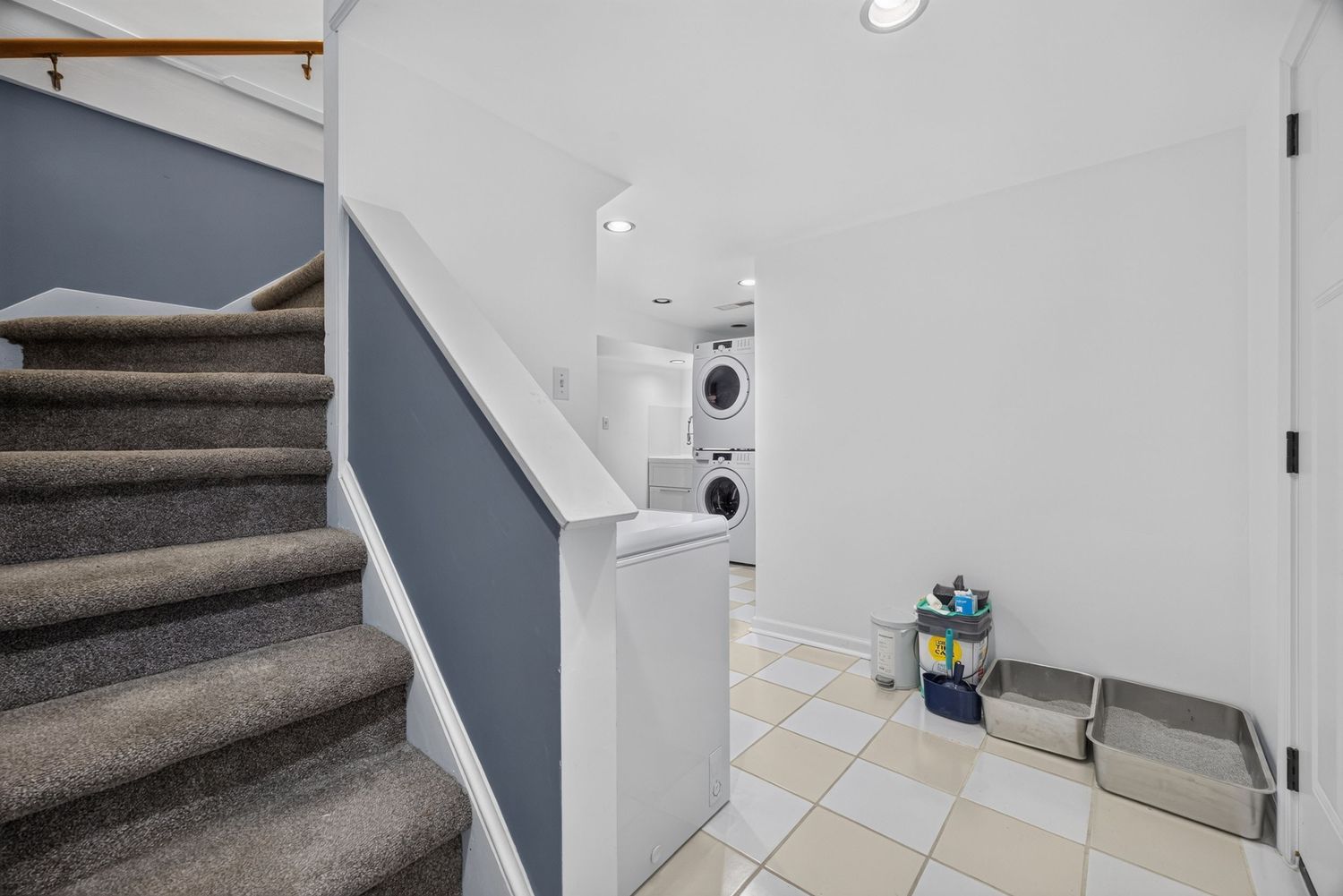Bright laundry room with stacked washer-dryer, white walls, tile floors, and carpeted staircase leading to upper level.
