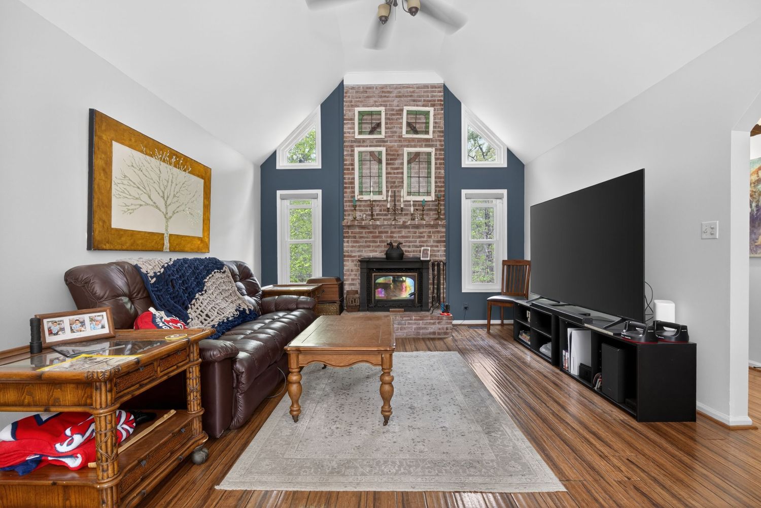 Cozy living room with vaulted ceilings, brick fireplace, leather sofa, hardwood floors, and large TV in a blue accent wall.