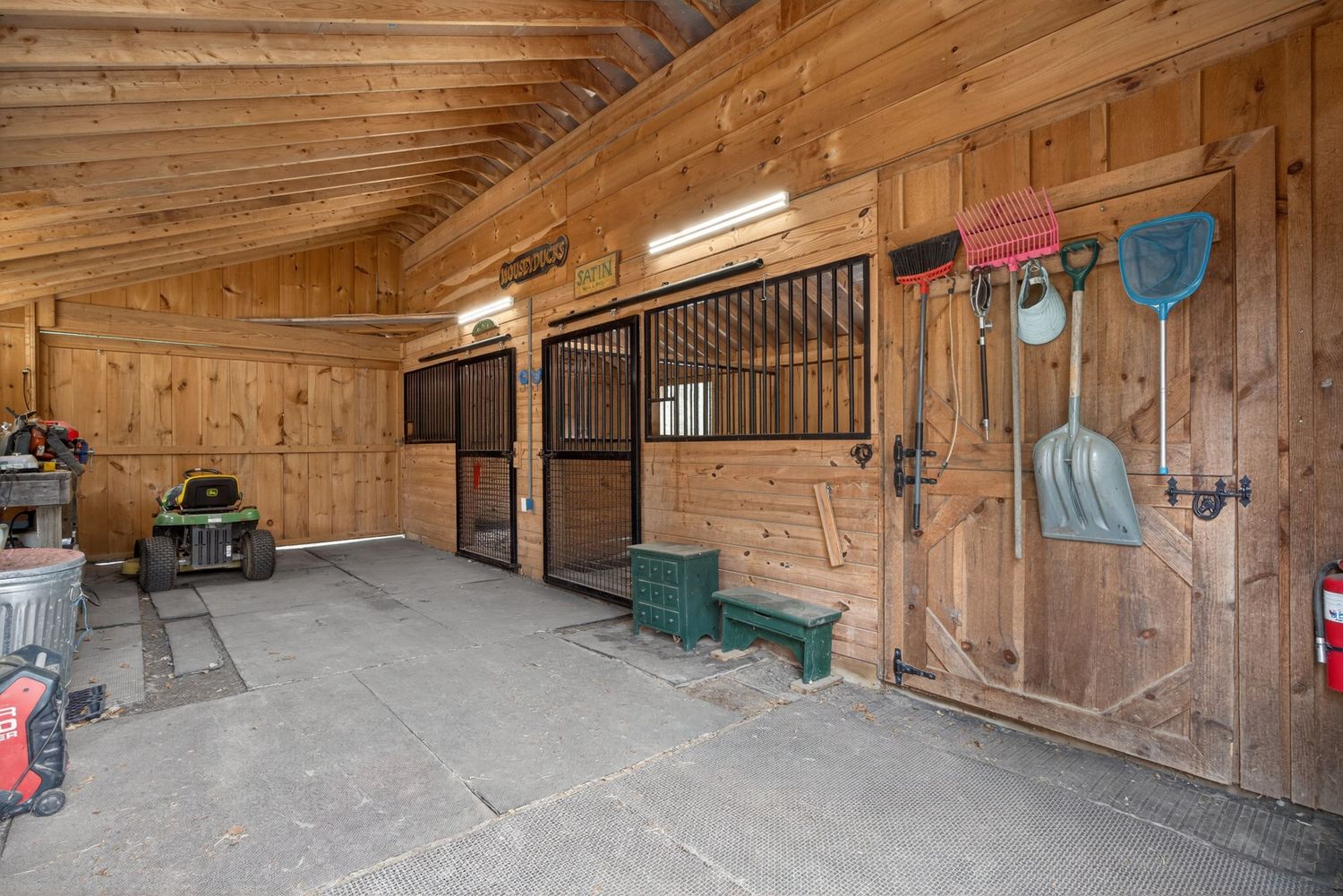 Rustic wooden horse barn interior with stall doors, concrete floor, and grooming tools hanging on the wall.