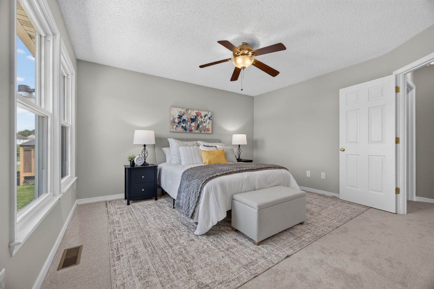 Spacious master bedroom with plush carpet, ceiling fan, neutral walls, large windows, and elegant bedding with a bench at the foot.