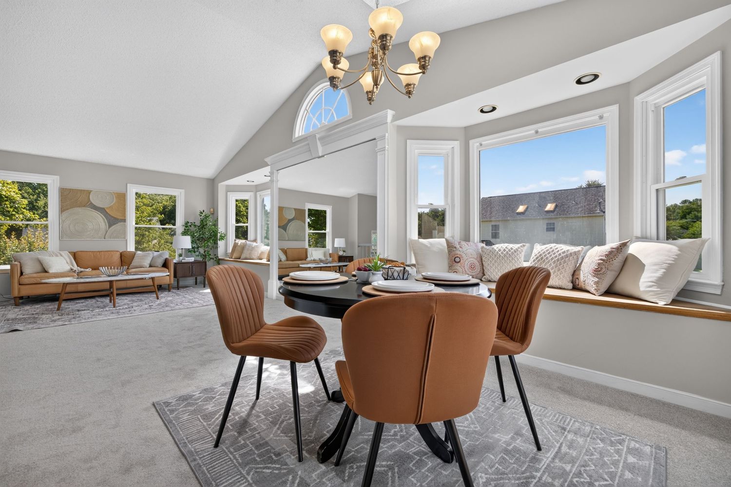 Bright dining room with round table, brown chairs, chandelier, vaulted ceiling, and bay windows overlooking a scenic view.