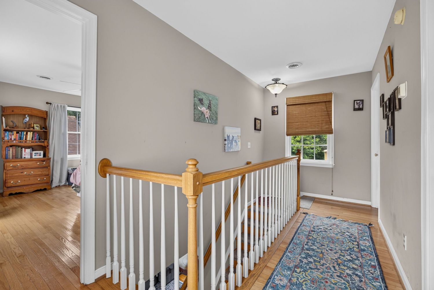 Bright upstairs hallway with hardwood floors, white stair railing, neutral walls, decorative rug, and natural light from a window.