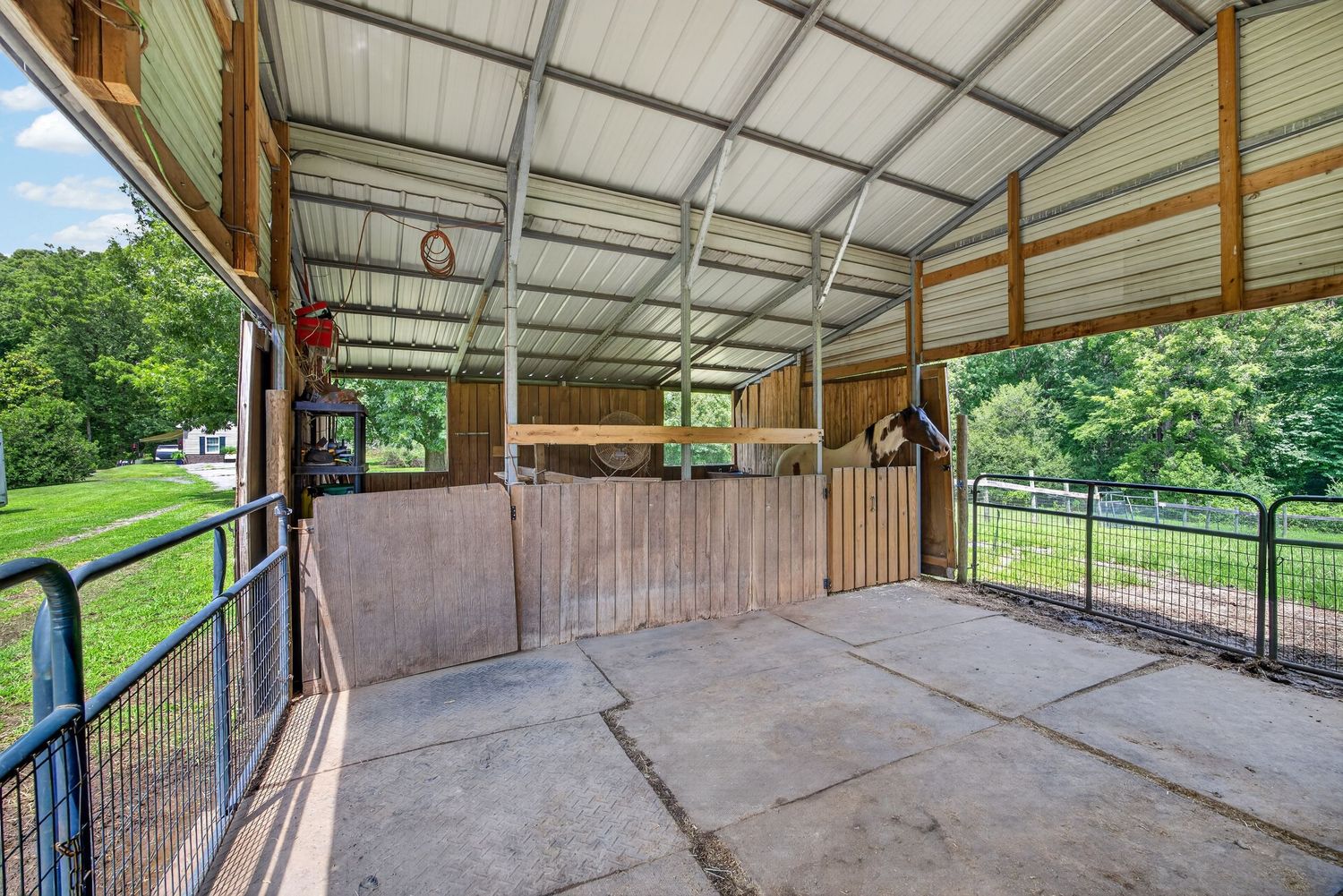 Open horse barn with metal roof, wooden stall dividers, concrete floor, and green pasture views on a sunny day.