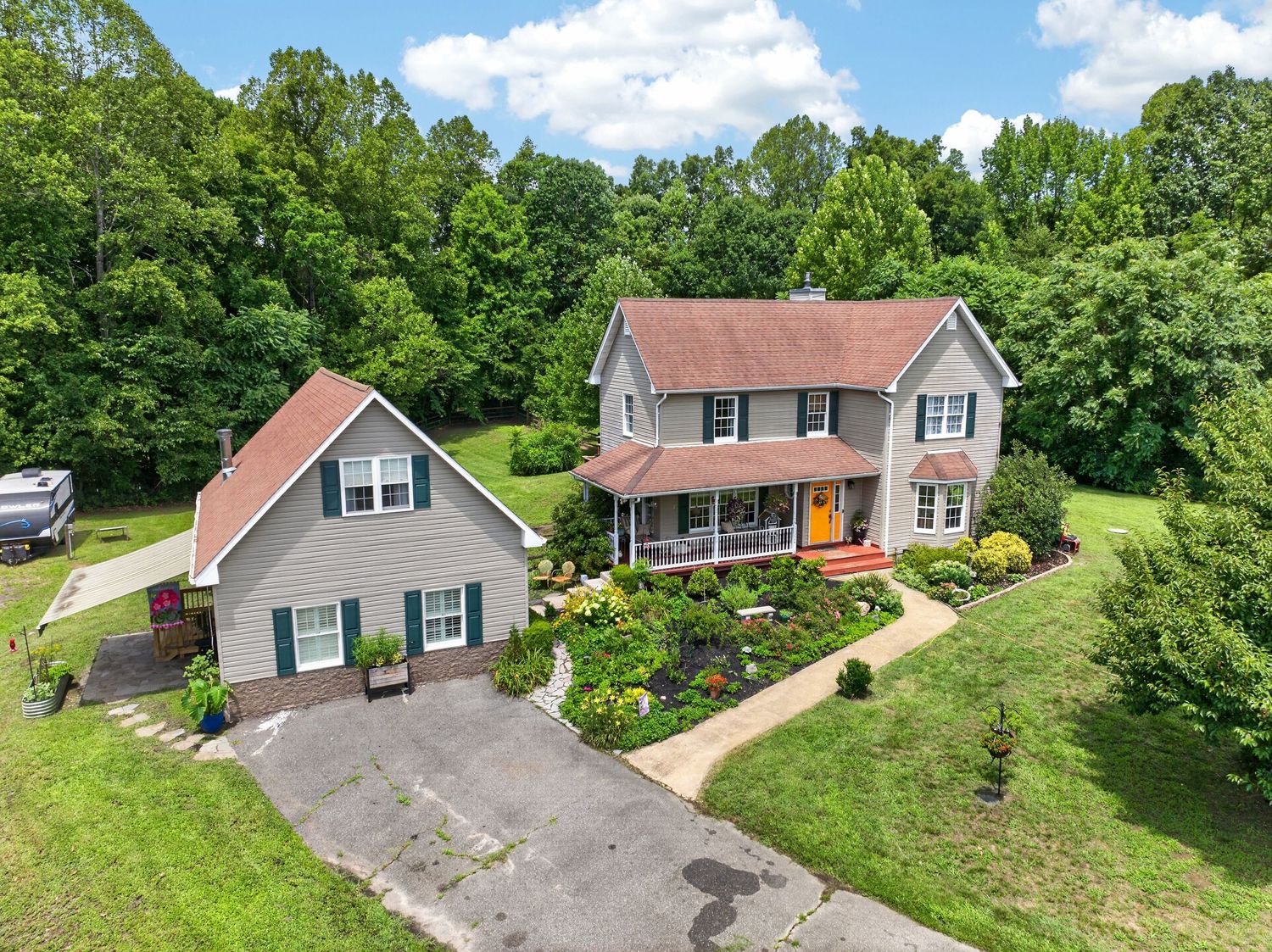 Aerial view of a two-story colonial home with gray siding, red roof, wraparound porch, and lush green landscaping.