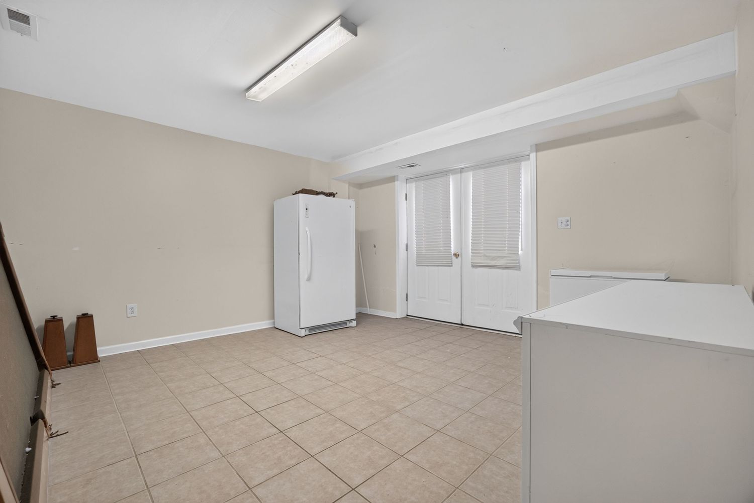 Bright empty room with white walls, tile flooring, fluorescent lighting, a white refrigerator, and glass panel doors.