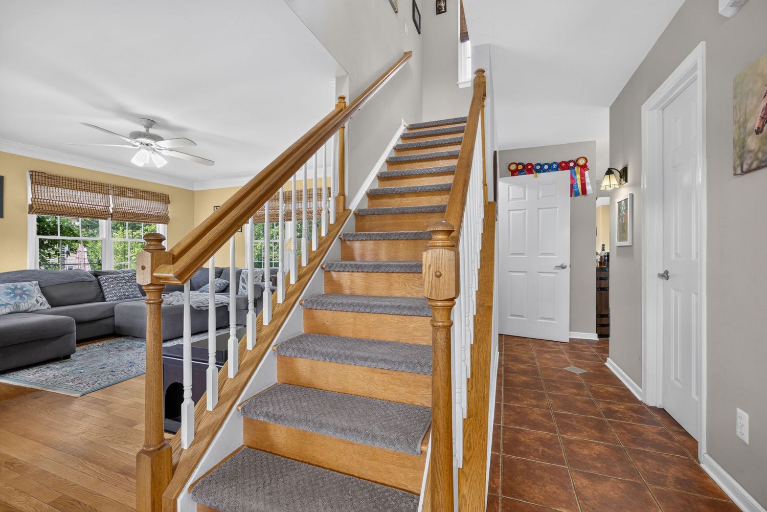 Bright home entryway featuring wooden staircase with gray carpet runner, ceiling fan, and tile flooring leading to living room.