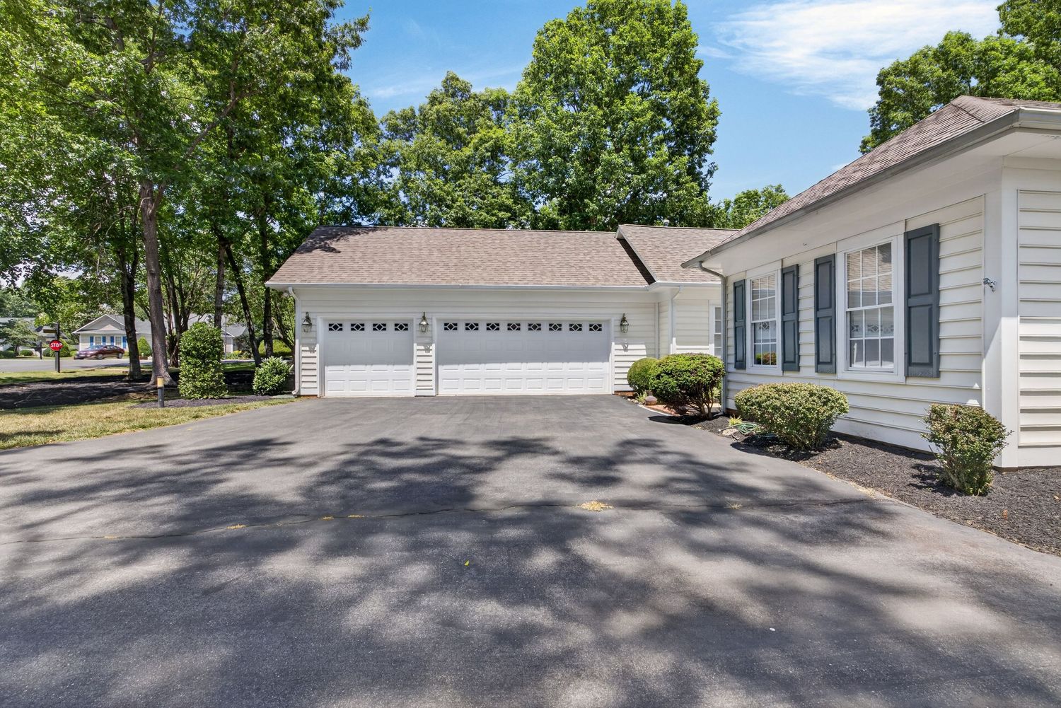White home with two-car garage, dark asphalt driveway, and mature trees in a well-maintained suburban neighborhood.