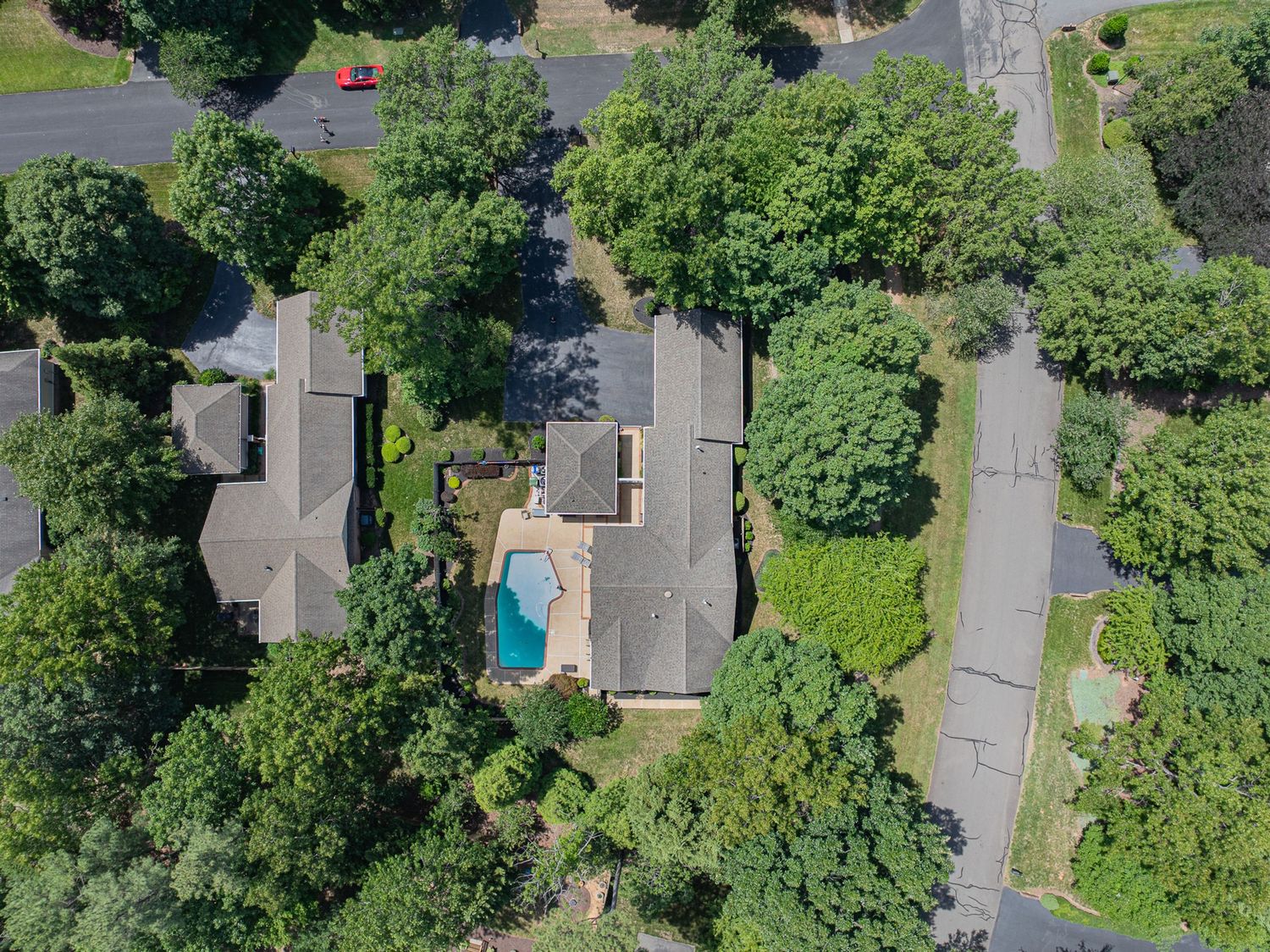 Aerial view of a suburban home with a backyard pool surrounded by lush green trees and a nearby road.