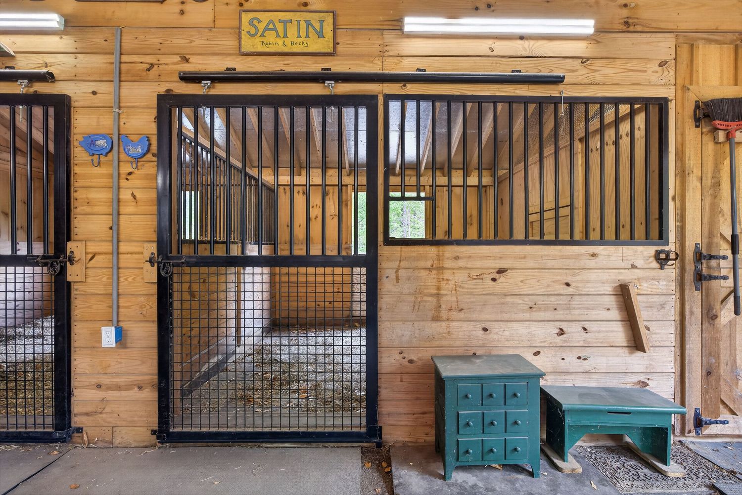 Horse stable interior with black metal stall gates, wooden walls, green cabinet, and a sign reading SATIN.