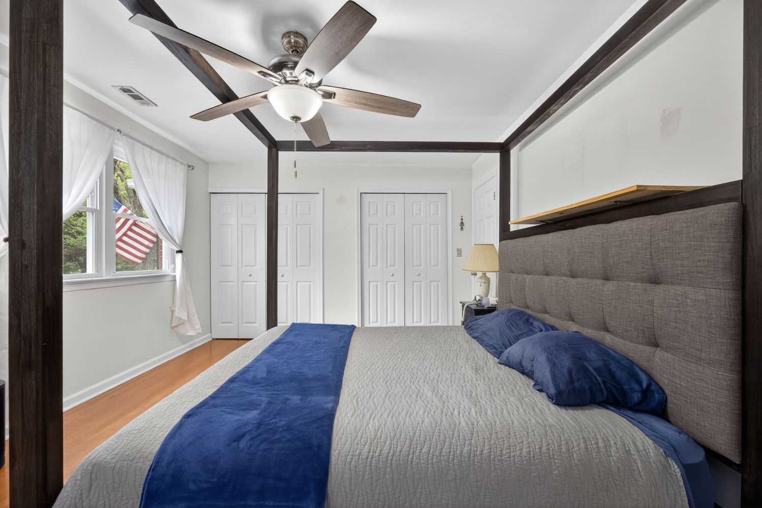 Cozy bedroom with gray upholstered bed, blue accents, ceiling fan, hardwood floors, and white closet doors.