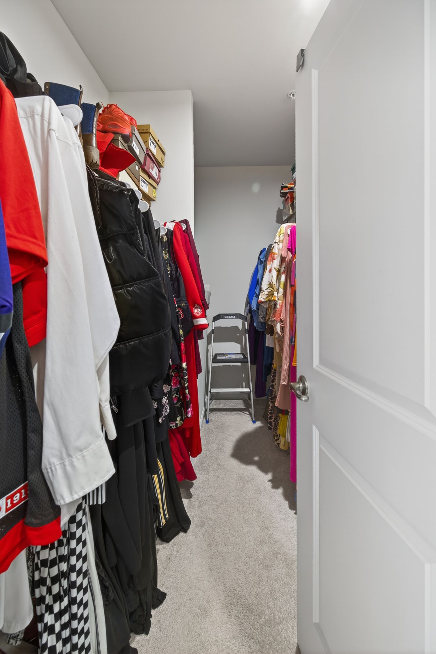 Narrow walk-in closet with colorful hanging clothes, racing gear, and a folding step ladder on carpeted floor.