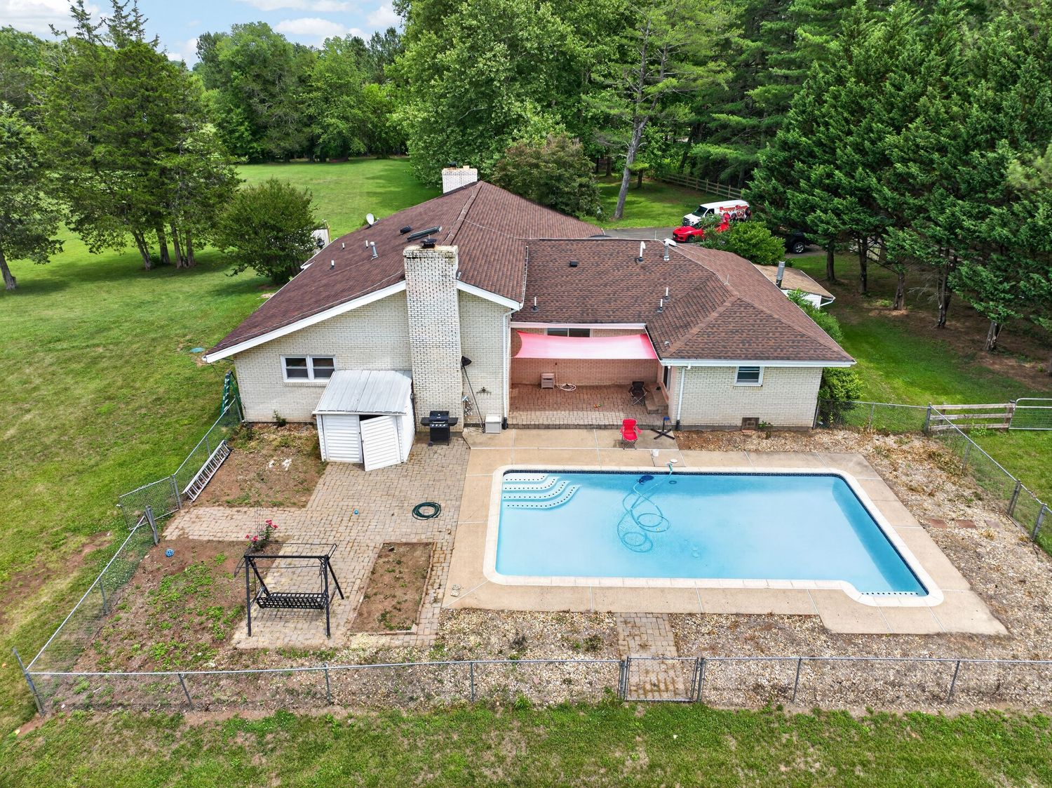 Aerial view of a ranch-style home with an in-ground pool, patio, and lush green surroundings.