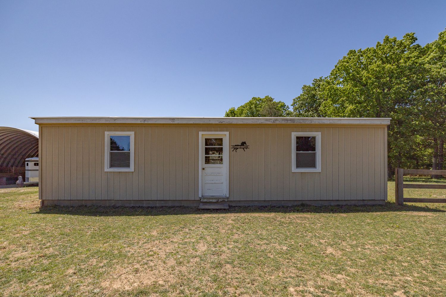 Small tan single-wide mobile home with white door and two windows on a rural property with green trees and open lawn.