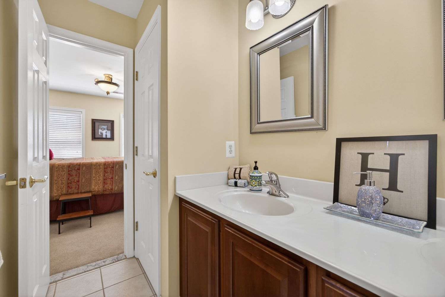 Elegant bathroom vanity with white countertop, cherry wood cabinets, framed mirror, and view into carpeted bedroom.