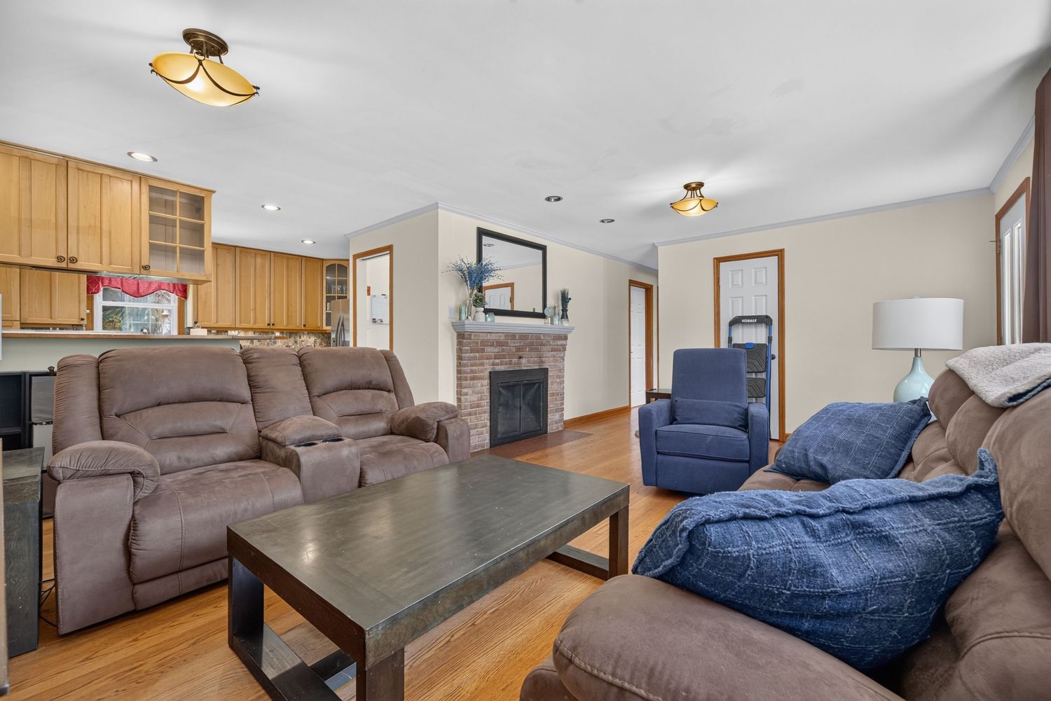 Cozy living room with brown reclining sofas, stone fireplace, hardwood floors, and open kitchen in background.