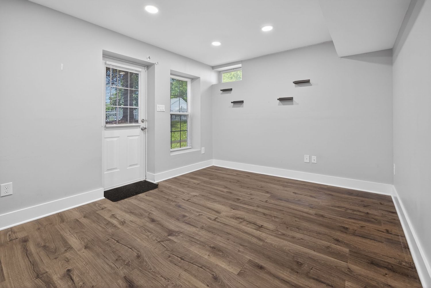 Bright empty room with dark hardwood floors, white walls, recessed lighting, and a white door with a window.