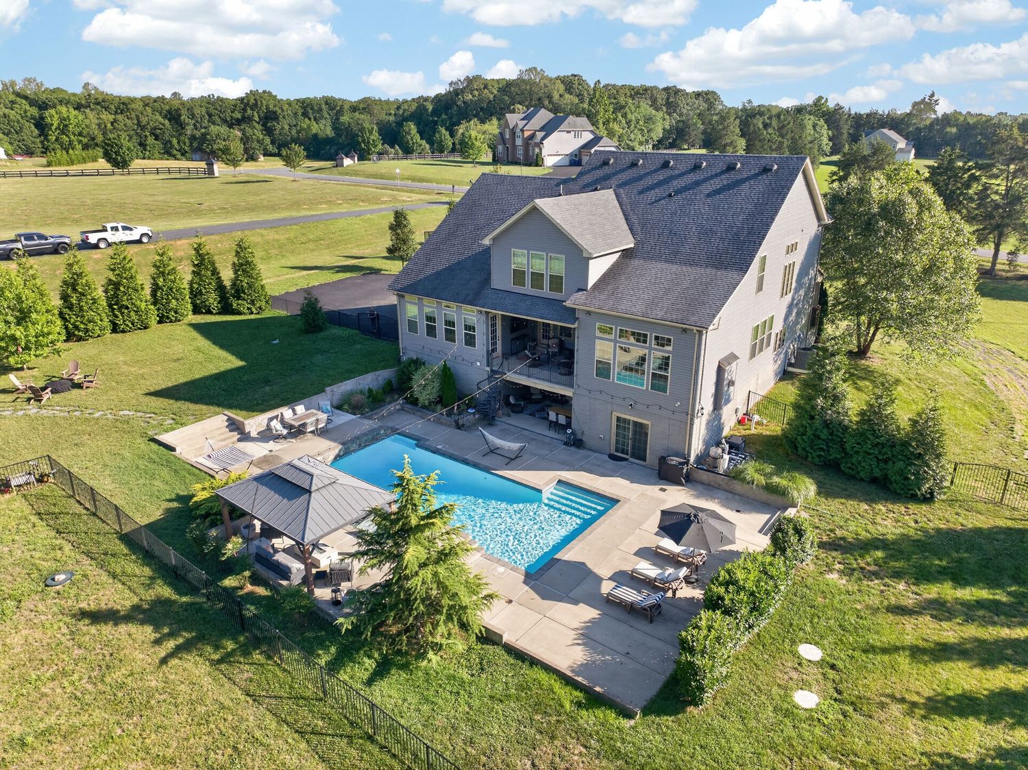 Aerial view of a large suburban home with a rectangular swimming pool, patio seating, and lush green lawn surroundings.