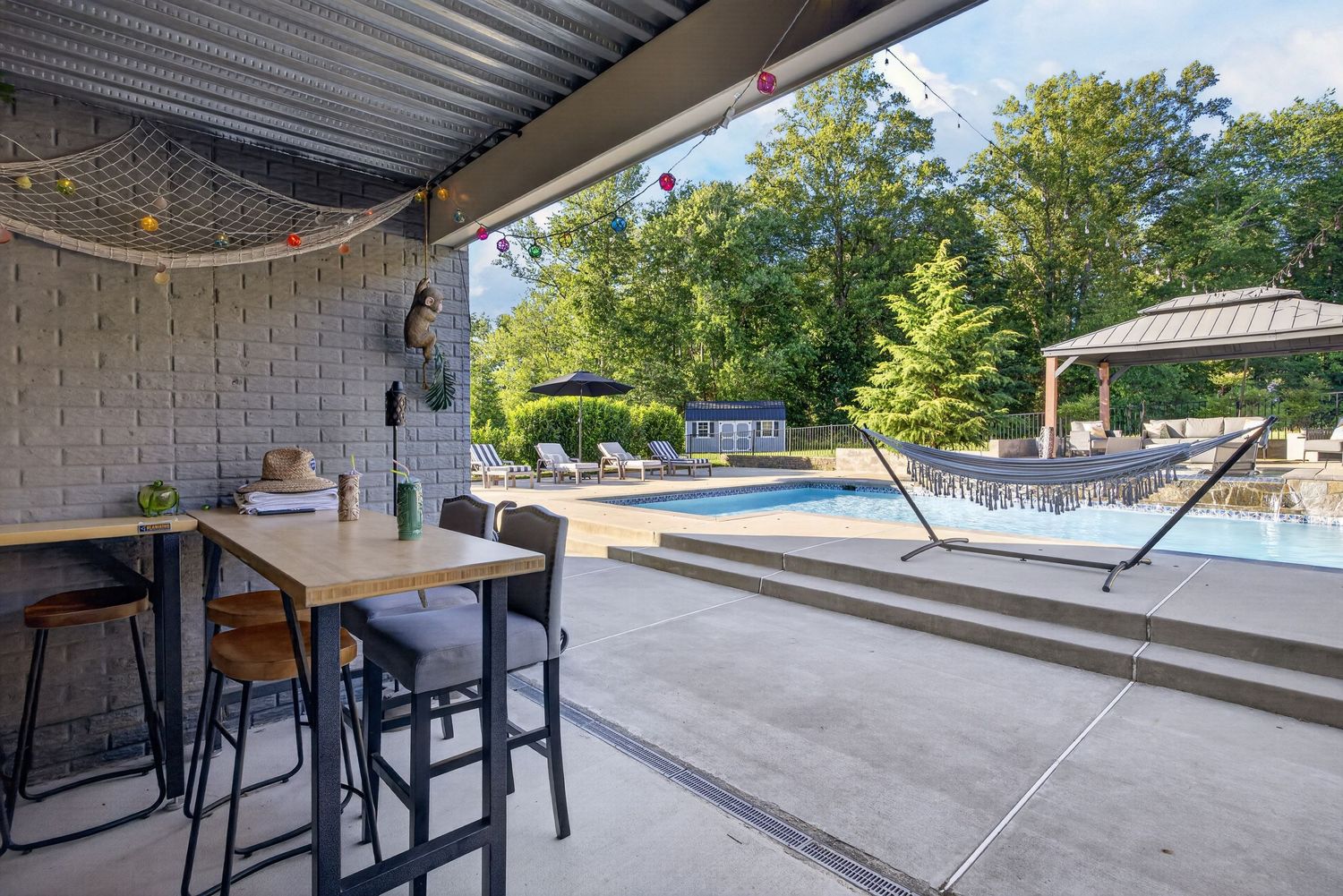 Covered patio with bar-height table, hammock, and sparkling outdoor pool surrounded by lush trees and a gazebo.