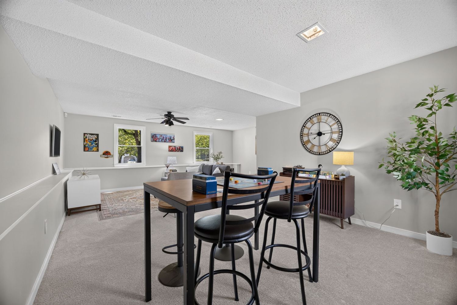 Modern dining area with bar-height table, black chairs, ceiling fan, and large clock in a bright carpeted living space.