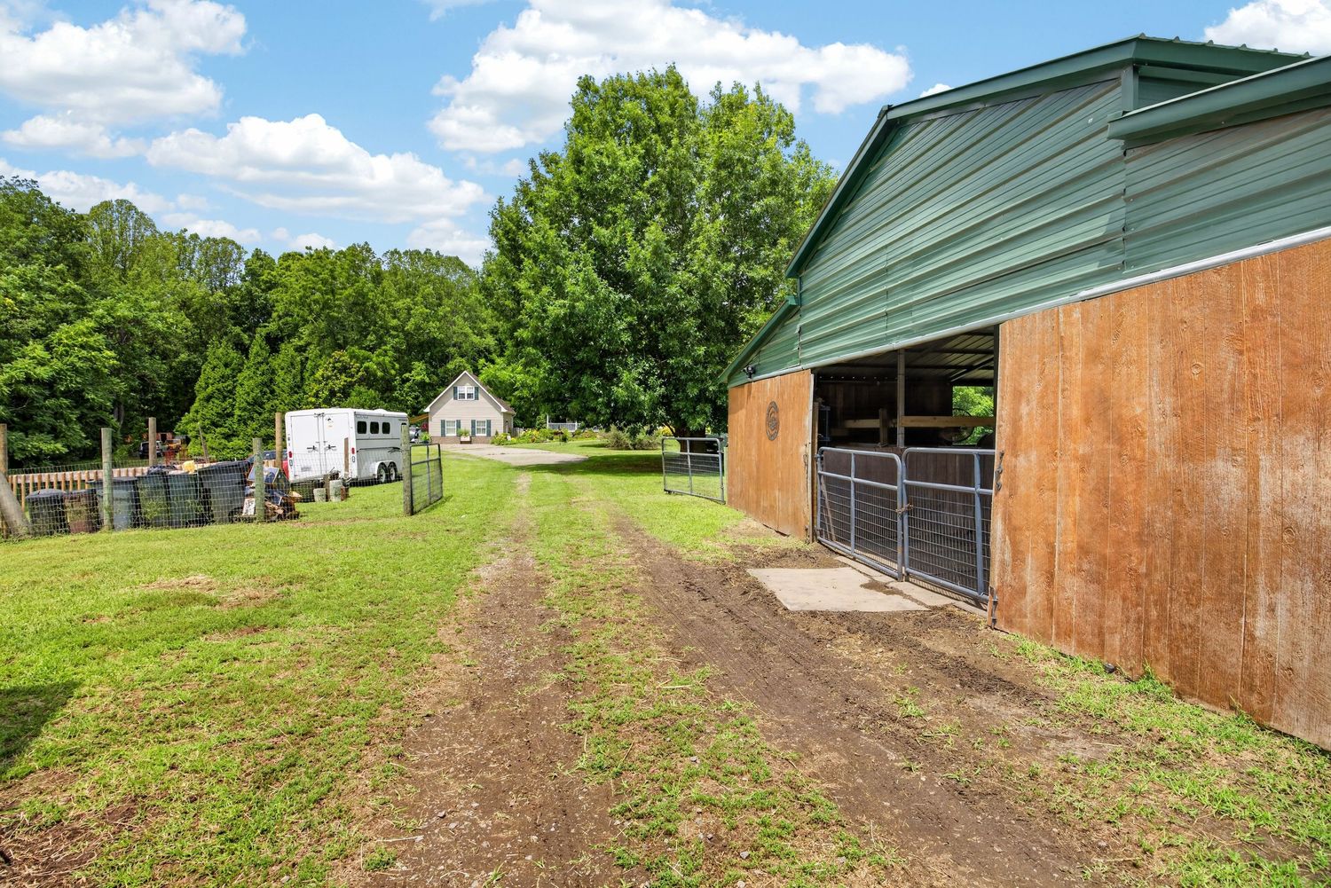 A rustic barn with green metal roof beside a dirt path on a lush farm property with trees and a white house in background.