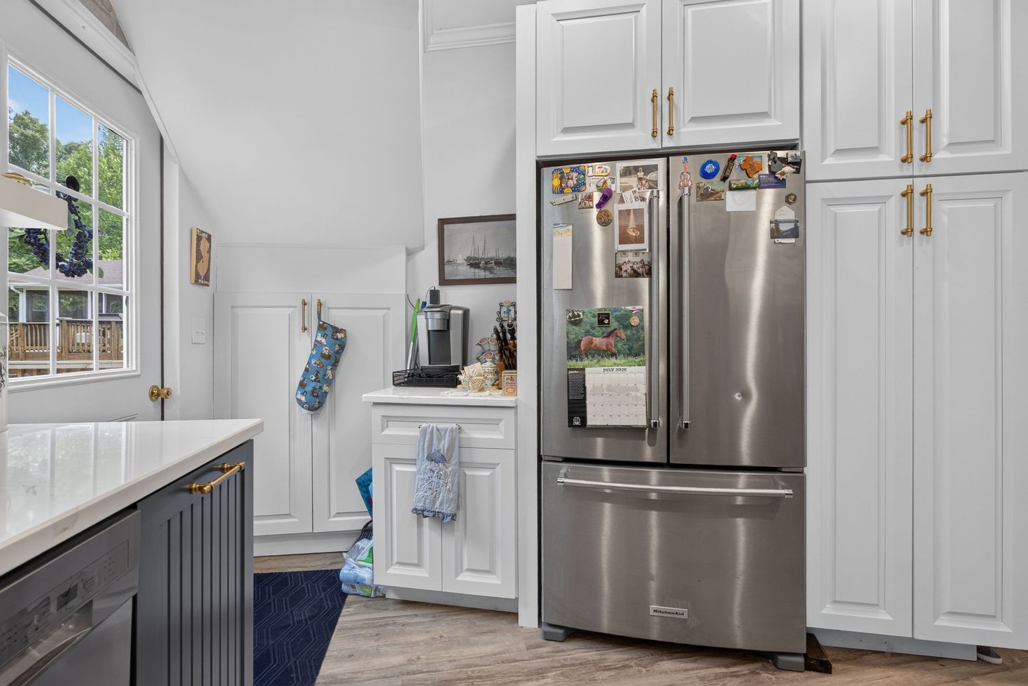 Modern kitchen with stainless steel French door refrigerator, white shaker cabinets with gold hardware, and hardwood floors.