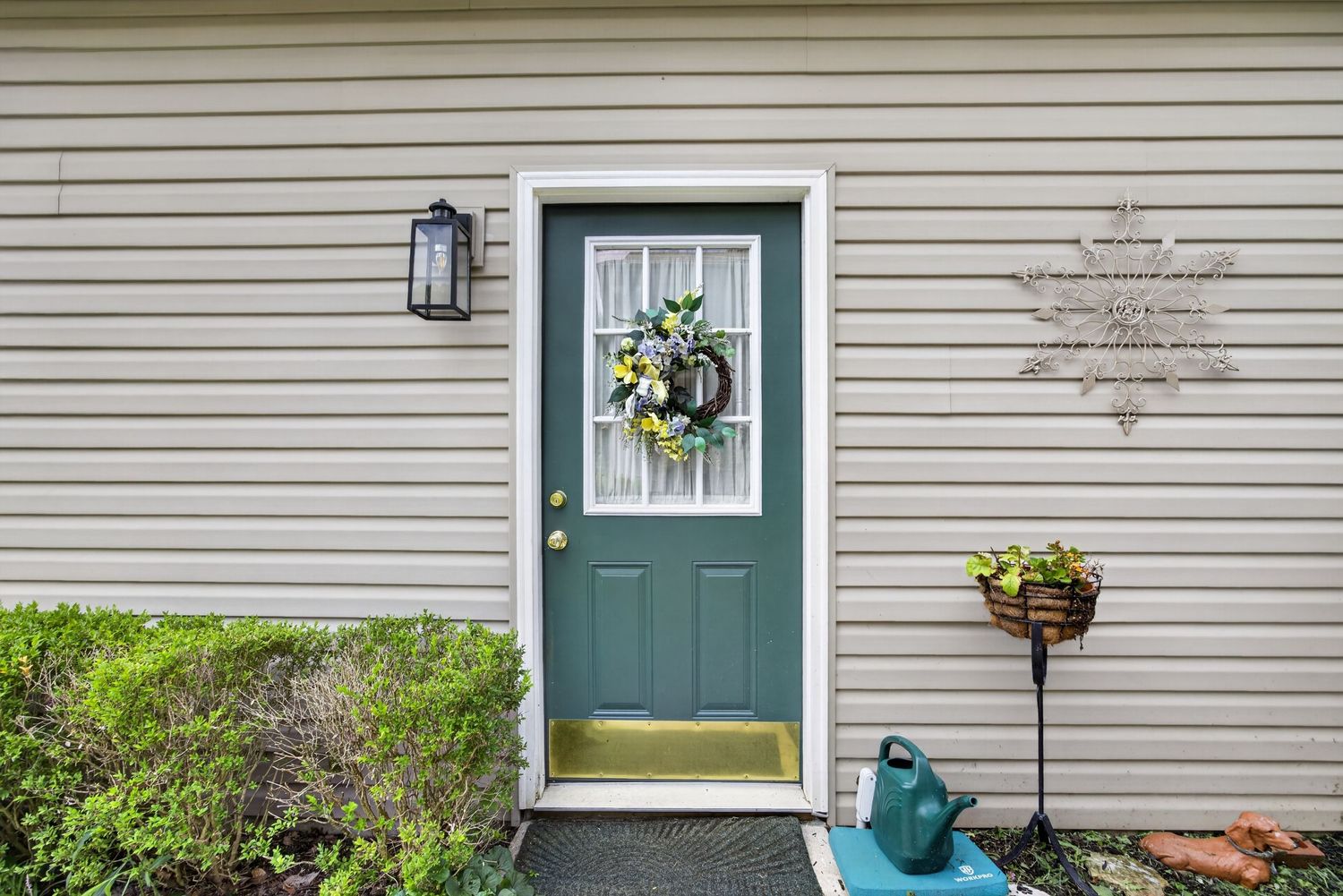 Teal front door with wreath, white trim, beige siding, wall lantern, and potted plants on a welcoming home entrance.