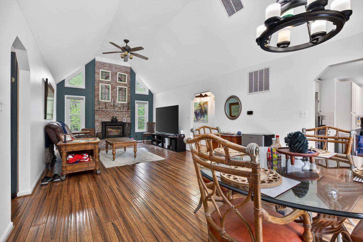 Open-concept living and dining room with hardwood floors, vaulted ceiling, ceiling fan, brick fireplace, and arched doorway.