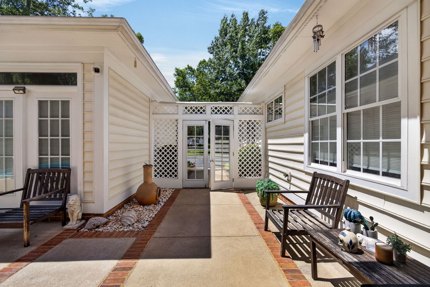 Charming white home exterior with a paved courtyard, lattice divider, benches, and lush greenery.