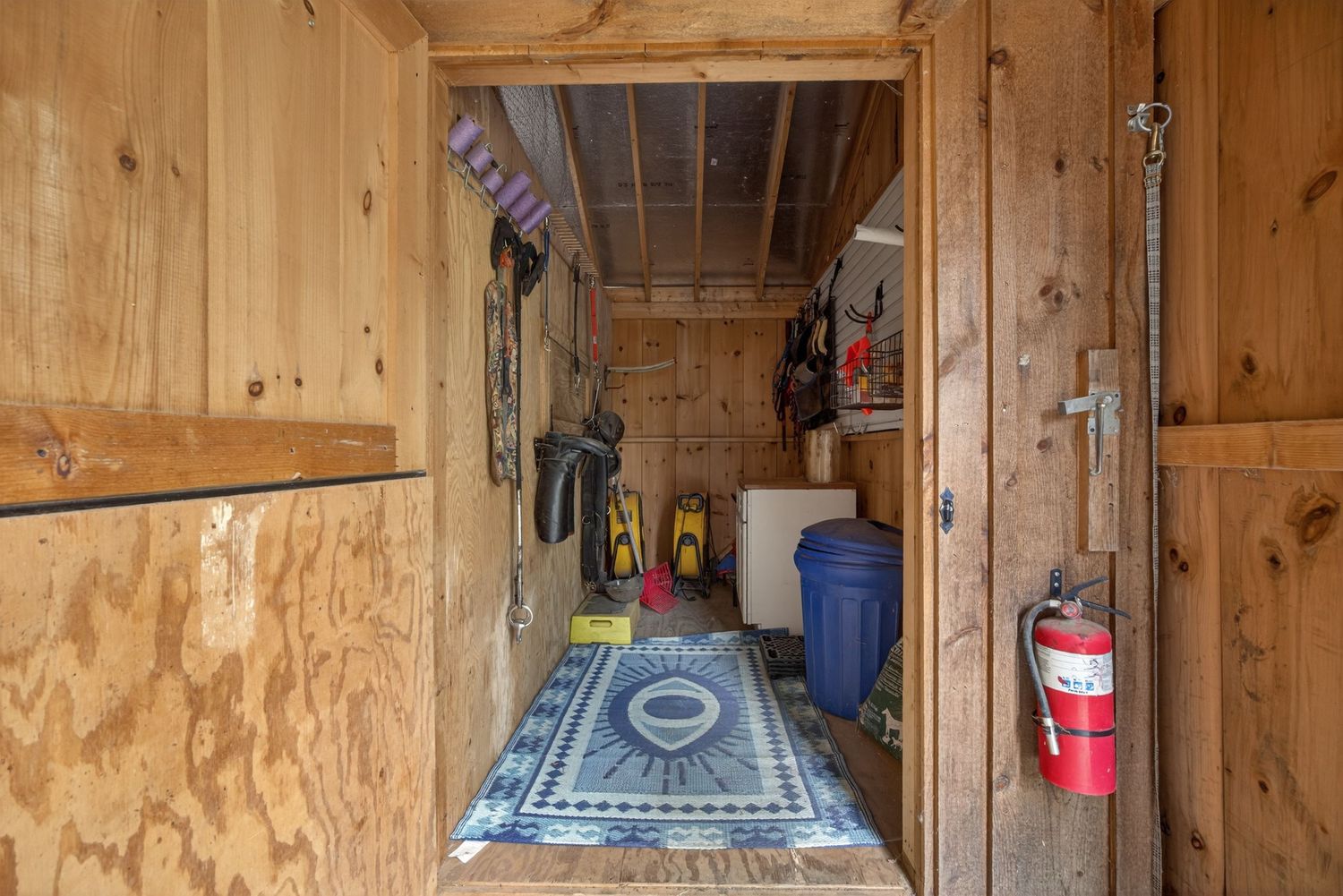Rustic wooden storage shed interior with tools, a blue trash can, area rug, and a red fire extinguisher by the door.