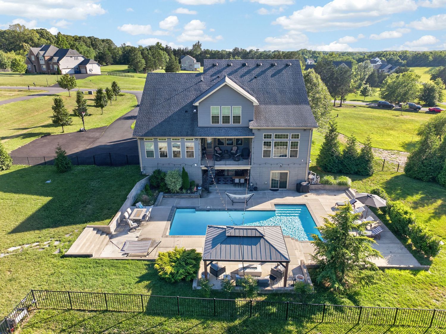 Aerial view of a large gray home with an in-ground pool, patio, gazebo, and lush green lawn in a rural setting.