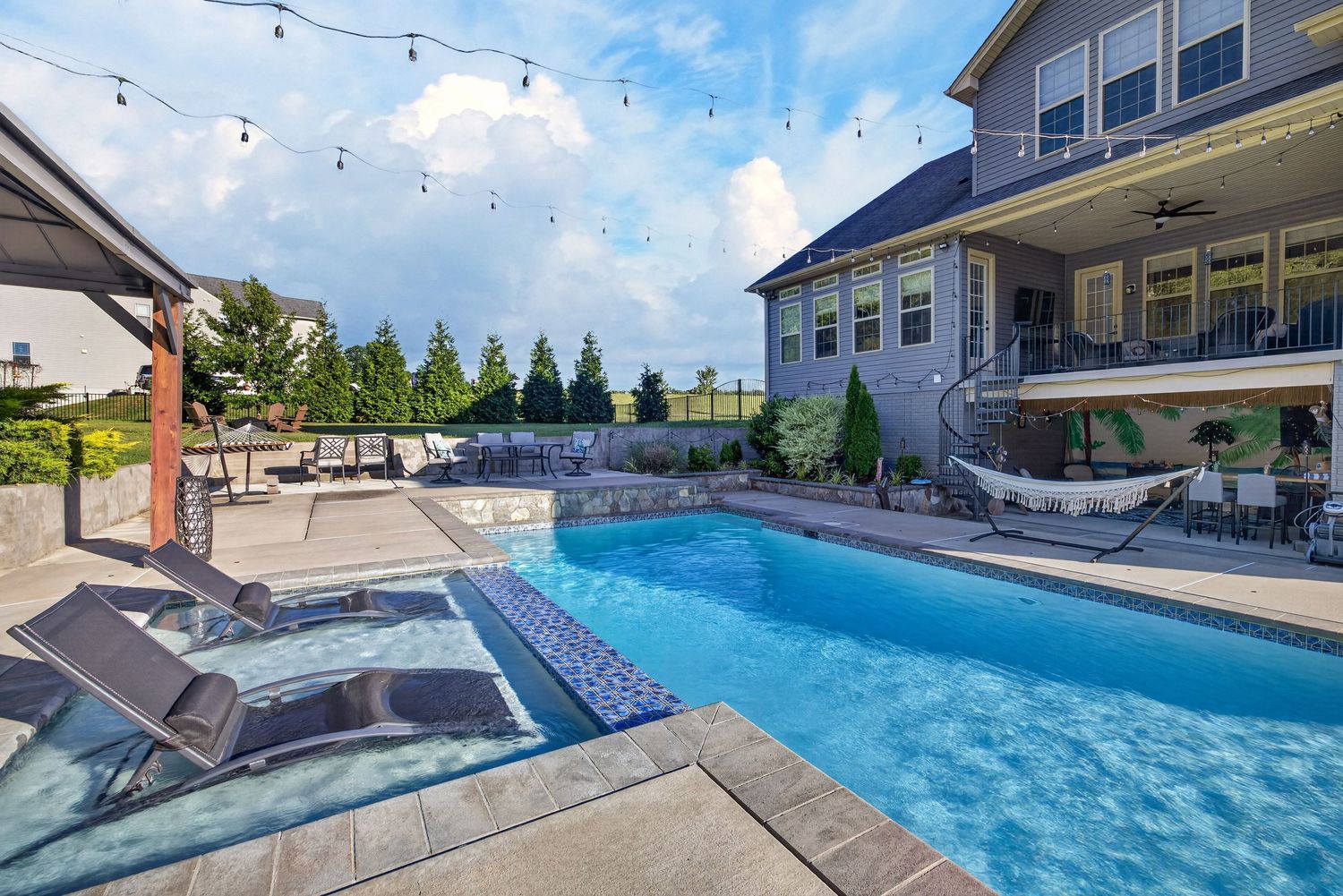 Luxury backyard pool with lounge chairs, string lights, and a large home with balcony on a sunny day.