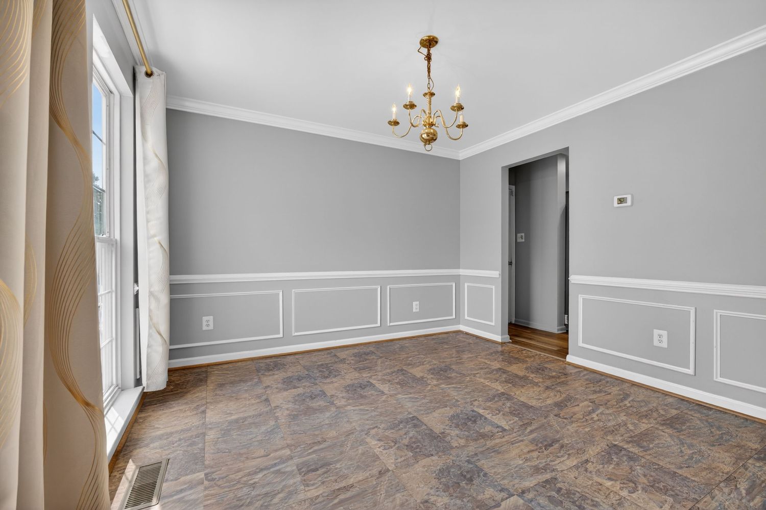 Empty dining room with gray wainscoting, dark stone tile floors, and a gold chandelier, bathed in natural light.