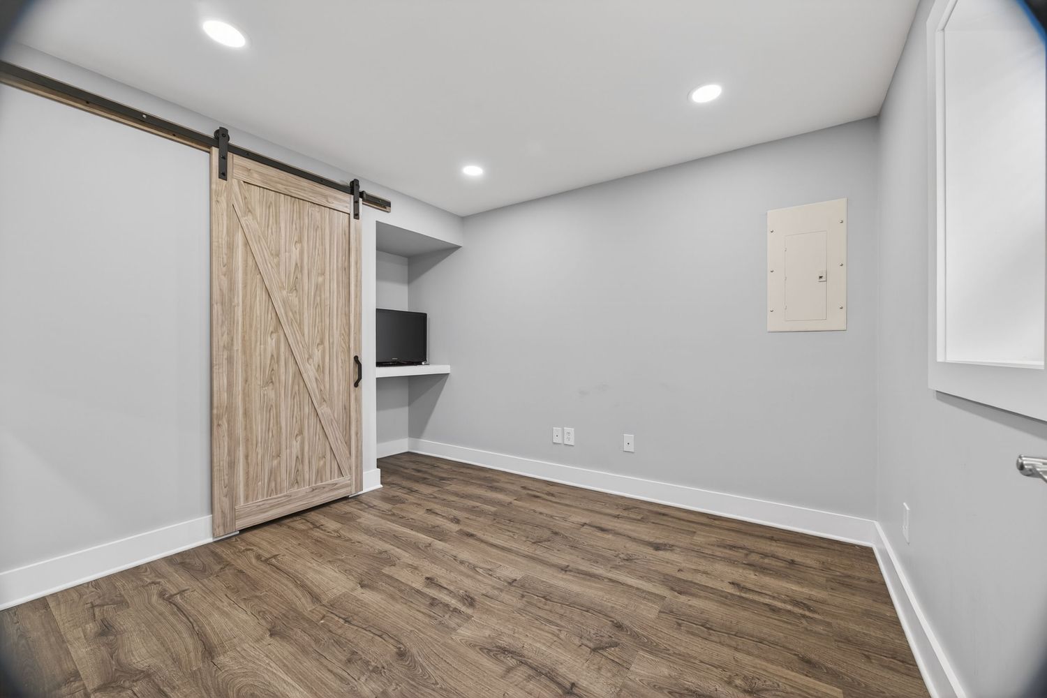 Modern basement room with barn door, built-in desk, hardwood floors, recessed lighting, and white walls.