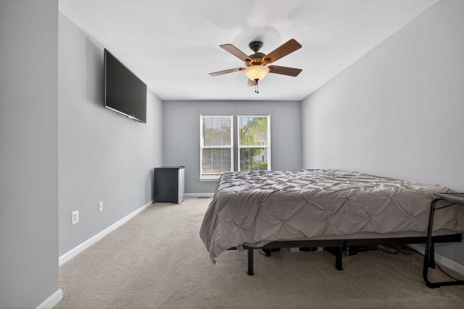 Spacious bedroom with gray carpet, ceiling fan, wall-mounted TV, and a large bed with gray bedding.