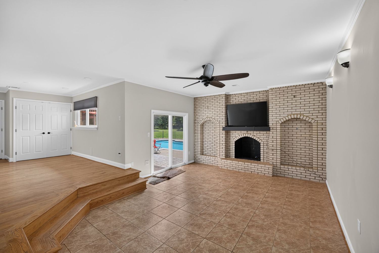 Spacious living room with brick fireplace, ceiling fan, tile floors, built-in shelves, and sliding doors leading to a pool.