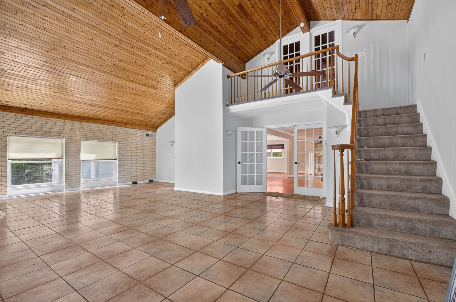 Spacious open-plan living area with tile floors, vaulted wood ceiling, staircase, and second-floor balcony in a bright home.