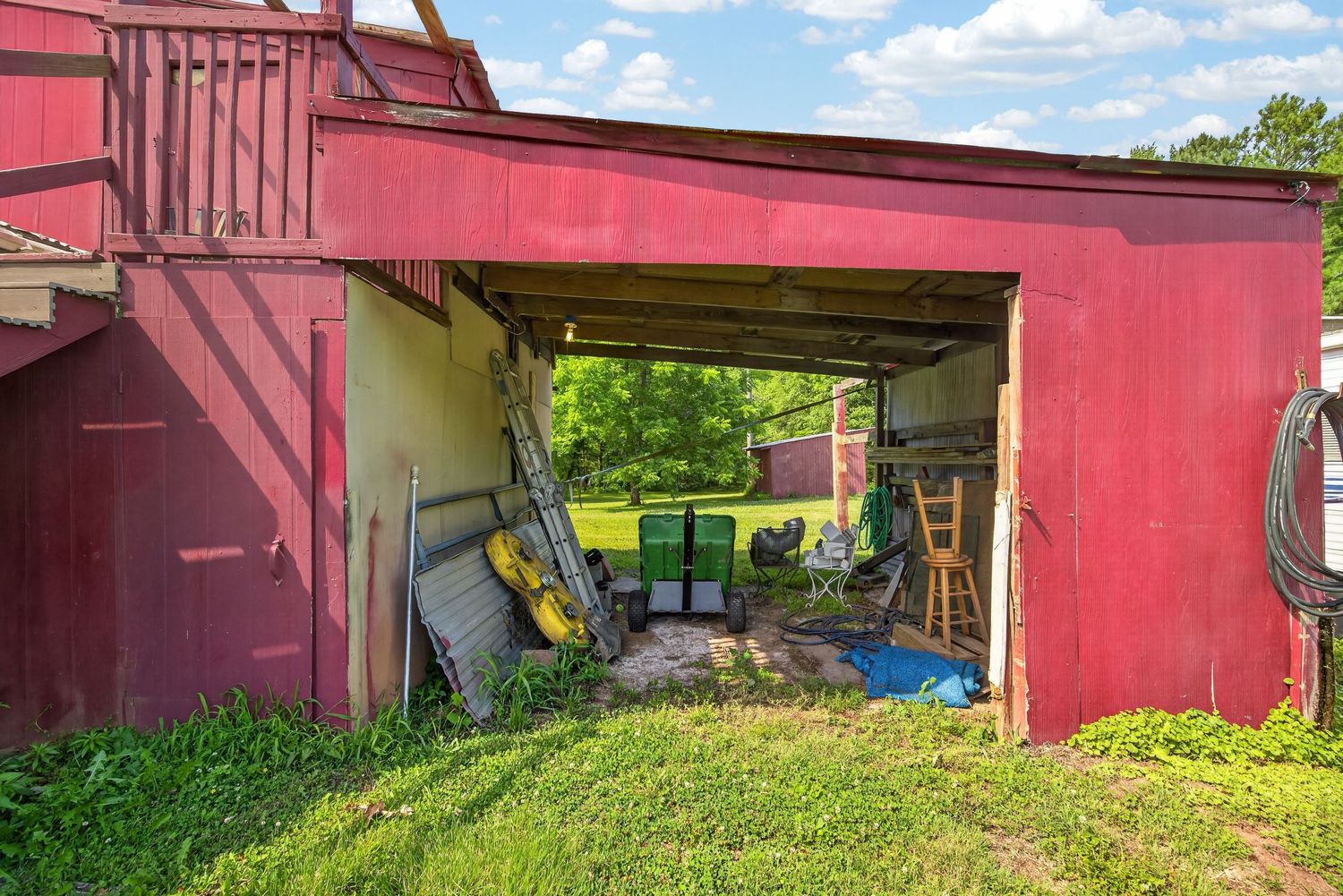 Red barn with open storage bay containing tools and equipment, surrounded by green grass and trees on a sunny day.