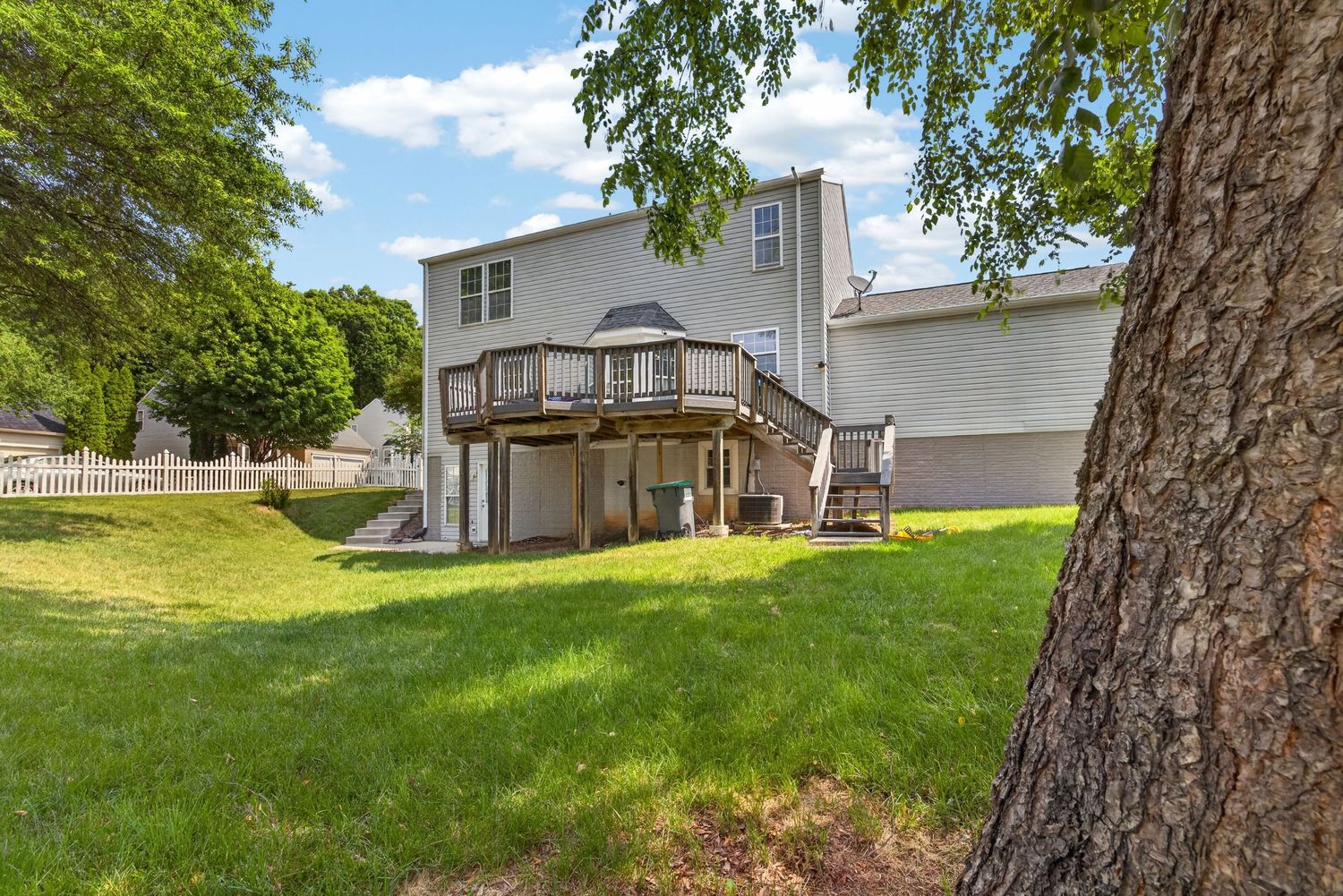 Two-story home with large wooden deck, lush green lawn, and mature trees in a suburban backyard setting.