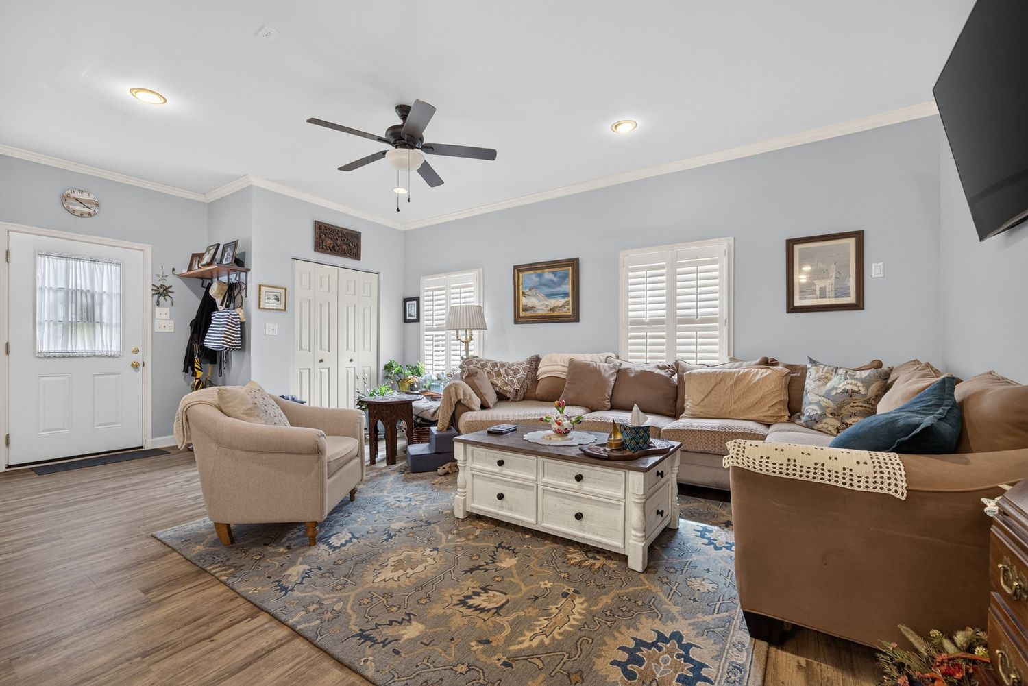 Bright living room with brown sectional sofa, ceiling fan, hardwood floors, and plantation shutters on windows.