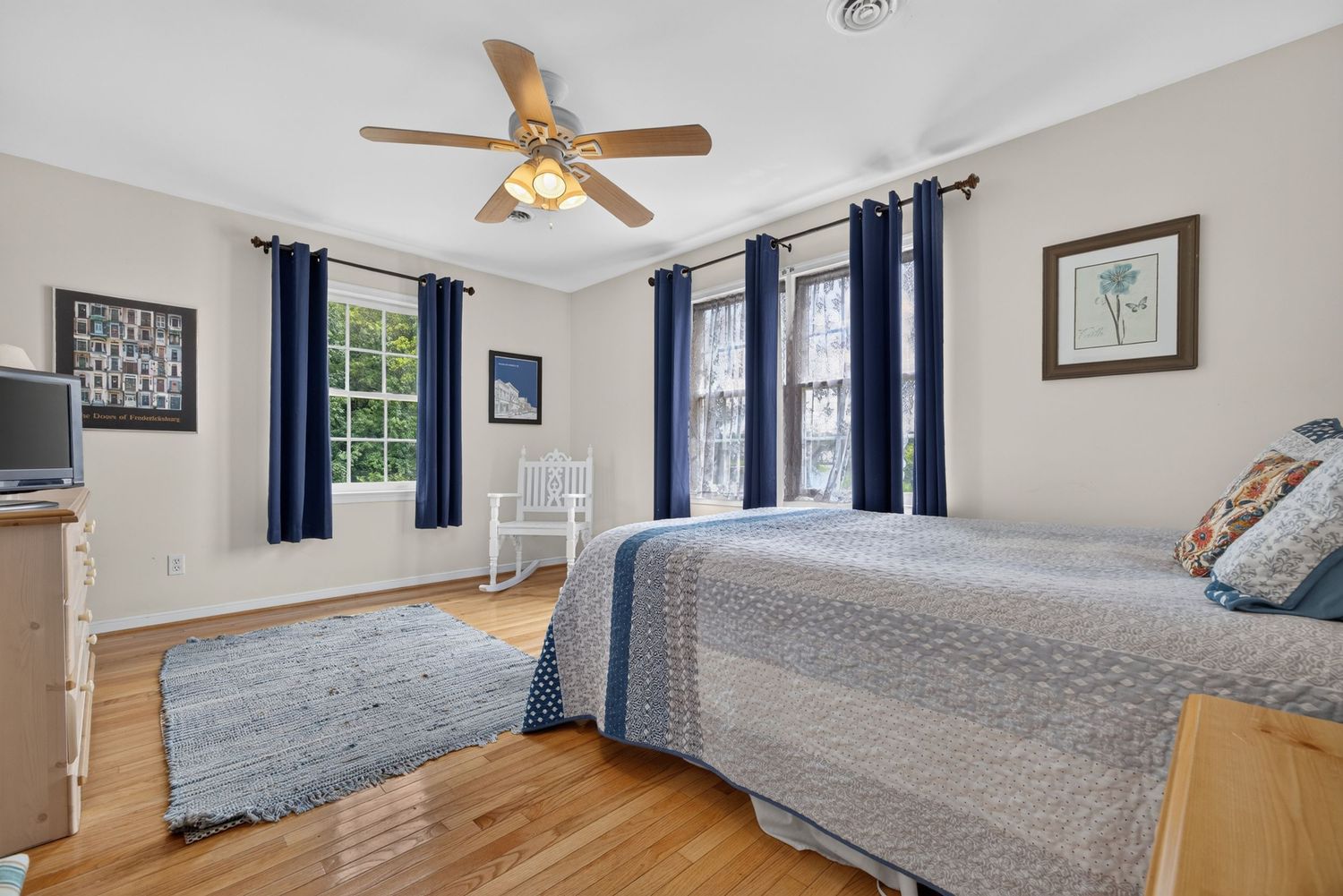 Bright bedroom with hardwood floors, blue curtains, ceiling fan, two beds with gray bedding, and a small blue rug.