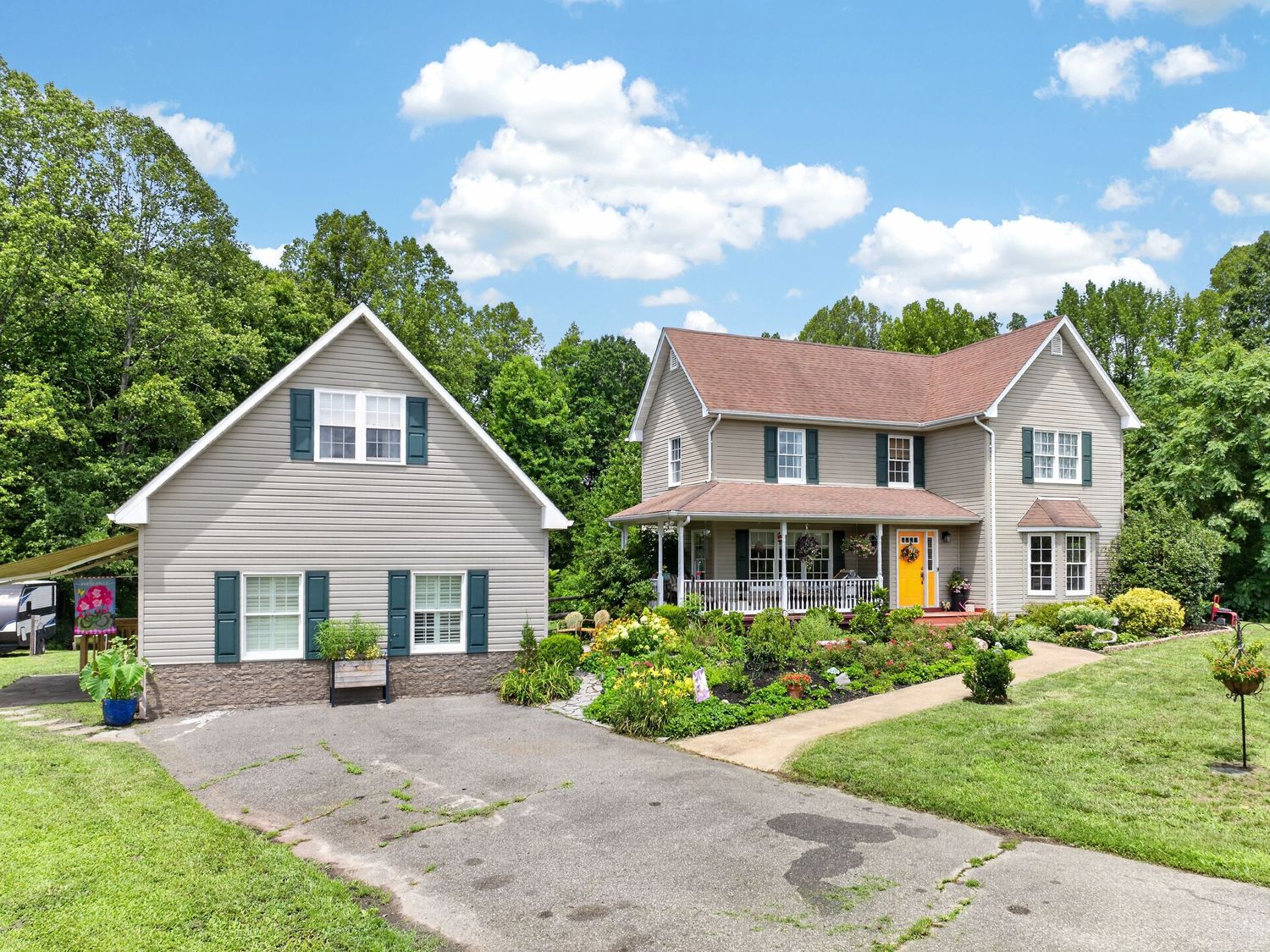 Two-story colonial home with gray siding, yellow door, covered porch, and lush green landscaping on a sunny day.