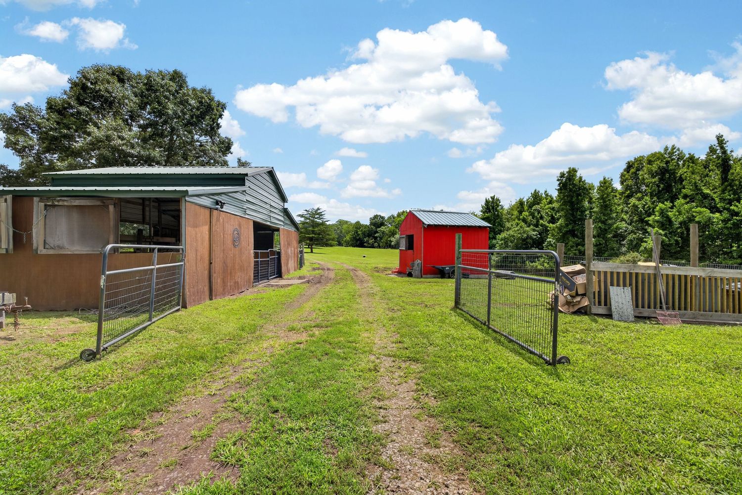 Rural farm property featuring a green-roofed stable, red shed, lush grass, metal gate, and tall trees under a blue sky.