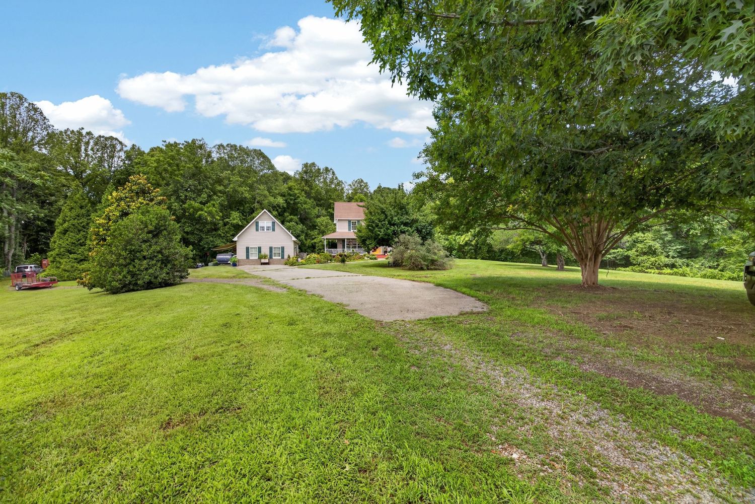 A white farmhouse sits at the end of a gravel driveway surrounded by lush green lawn and mature trees on a sunny day.