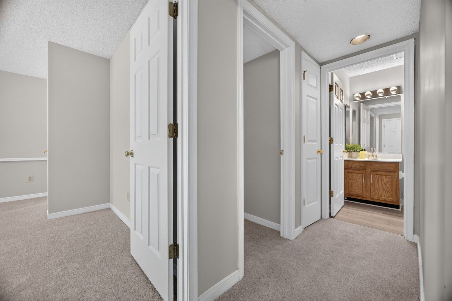 Carpeted hallway with white six-panel doors leading to a bathroom with wood vanity and mirror lighting.