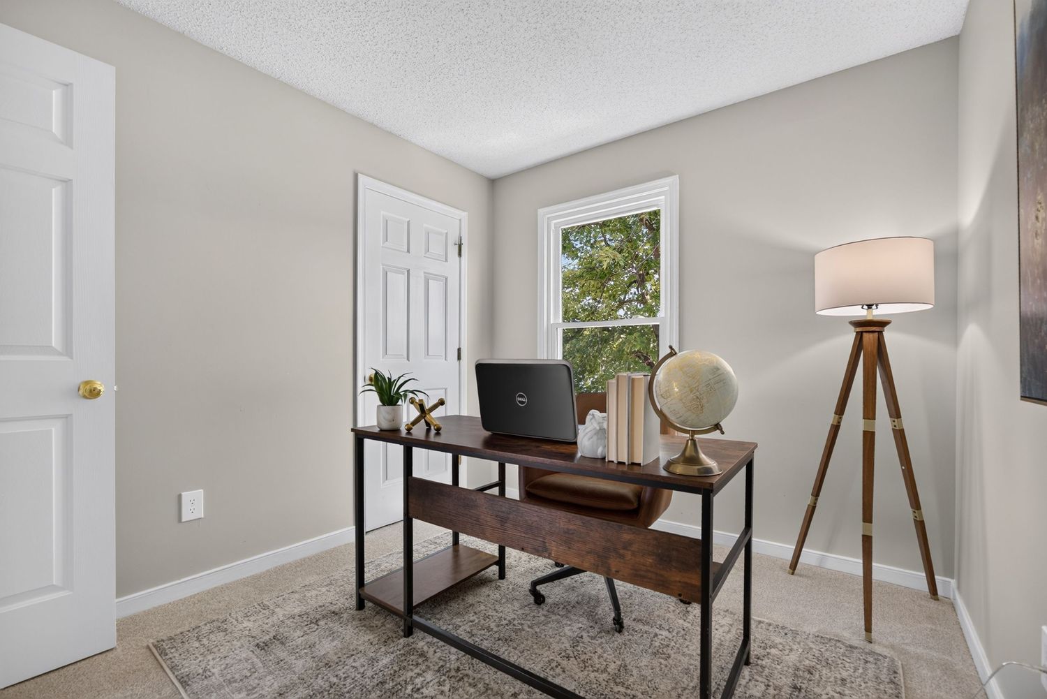 A cozy home office with a rustic wooden desk, laptop, globe, and tripod floor lamp on a patterned rug.